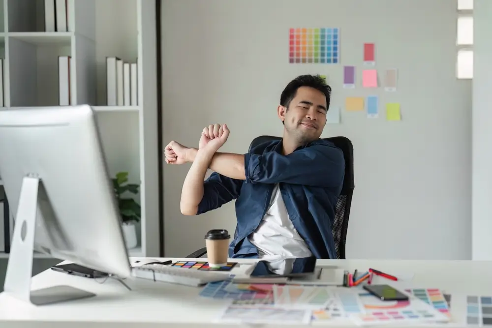 Graphic Designer Faire une pause pour étirer les muscles dans un environnement de bureau moderne avec des outils créatifs et des nuances de couleurs.