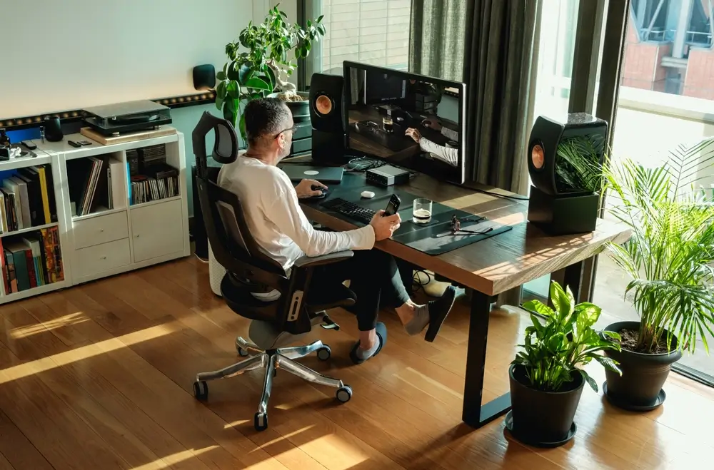 Vue de dessous d'homme d'âge mûr travaillant sur son ordinateur à la maison par une fenêtre sur un lieu de travail moderne dans une pièce décorée de plantes vertes en pot.